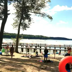 Słoneczny dzień nad jeziorem. Ludzie odpoczywają na plaży i molo, ciesząc się wakacjami. W tle las i błękitne niebo.