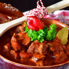 Gulasz w glinianej misie, ozdobiony papryczką i natką, z drewnianą łyżką pełną pieprzu i precelkami w tle. Idealny na chłodne wieczory, smakuje jak u babci.