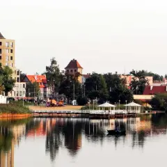 Widok na jezioro z łodzią, pomostem z altanami i budynkami w tle.