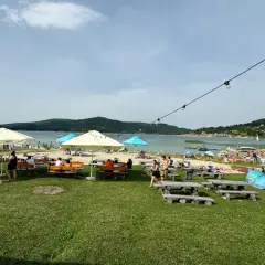 Plaża nad jeziorem z ludźmi, parasolami, stolikami i ławkami w słoneczny dzień.