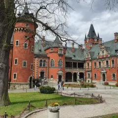 Pławniowice atrakcje: ceglany zamek z wieżą i zegarem, dziedziniec z drzewem i ludźmi spacerującymi po okolicy.