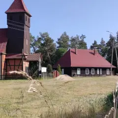 Drewniana wieża kościelna i dom z czerwonym dachem w Starowej Górze, otoczone polami i drzewami.
