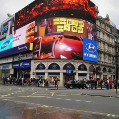 Tętniący życiem Piccadilly Circus w Londynie z neonowymi reklamami, w tym Samsung, McDonald's i Hyundai. Tłum ludzi na chodniku.