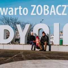 Rodzina pozuje przy napisie GDYNIA. W tle nowoczesne budynki, idealne na wakacyjne gdynia atrakcje dla dorosłych.