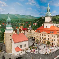 Miniaturowa panorama miasta z wieżami, ratuszem i placem pełnym postaci. W tle wzgórza i lasy, jakby z krainy jagniątków.