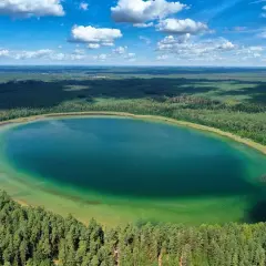 Widok z lotu ptaka na owalne, turkusowe jezioro otoczone gęstym lasem sosnowym.