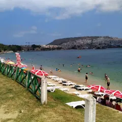 Plaża z leżakami, parasolami i ludźmi kąpiącymi się w morzu, w tle miasto na wzgórzu.
