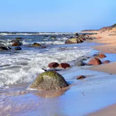 Kamienista plaża w Dębinie, z falami rozbijającymi się o brzeg i dwiema osobami spacerującymi po piasku. Dębina atrakcje przyrodnicze zachwycają.