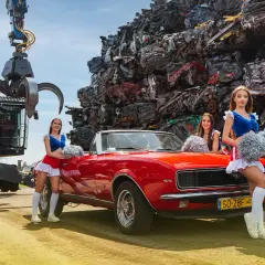 Trzy cheerleaderki w czerwono-niebieskich strojach stoją obok czerwonego kabrioletu na tle złomowiska.