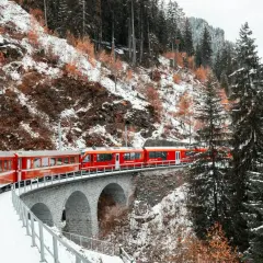 Czerwony pociąg Glacier Express przejeżdża przez kamienny most w zimowym, zaśnieżonym krajobrazie górskim.