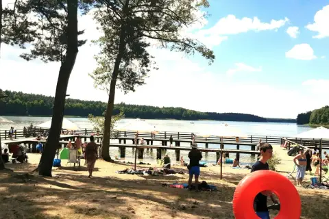 Słoneczny dzień nad jeziorem. Ludzie odpoczywają na plaży i molo, ciesząc się wakacjami. W tle las i błękitne niebo.