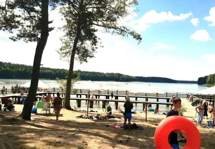 Słoneczny dzień nad jeziorem. Ludzie odpoczywają na plaży i molo, ciesząc się wakacjami. W tle las i błękitne niebo.