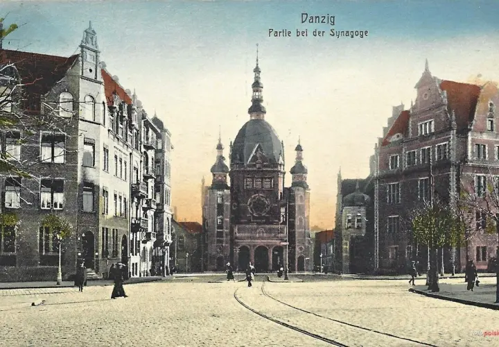 Widok na plac z imponującą synagogą w Gdańsku. Wokół kamienice, tory tramwajowe i kilka postaci spacerujących po bruku.