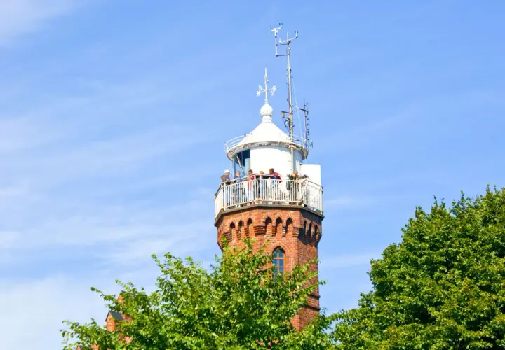 Widok na ceglaną latarnię morską w Ustce z tarasem widokowym, na którym stoją ludzie. W tle błękitne niebo i zielone drzewa.