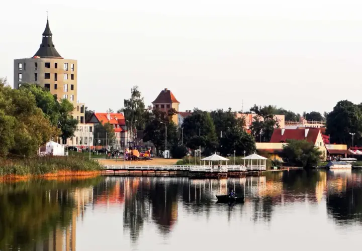 Widok na jezioro z łodzią, pomostem z altanami i budynkami w tle.