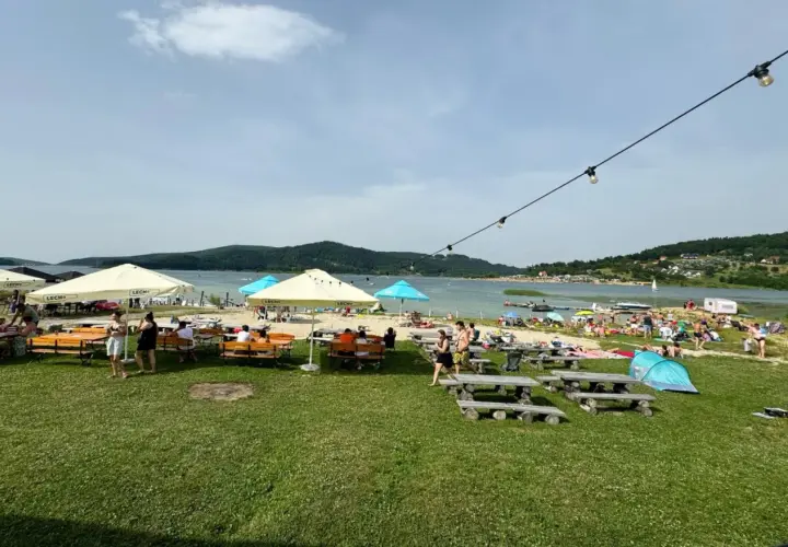 Plaża nad jeziorem z ludźmi, parasolami, stolikami i ławkami w słoneczny dzień.