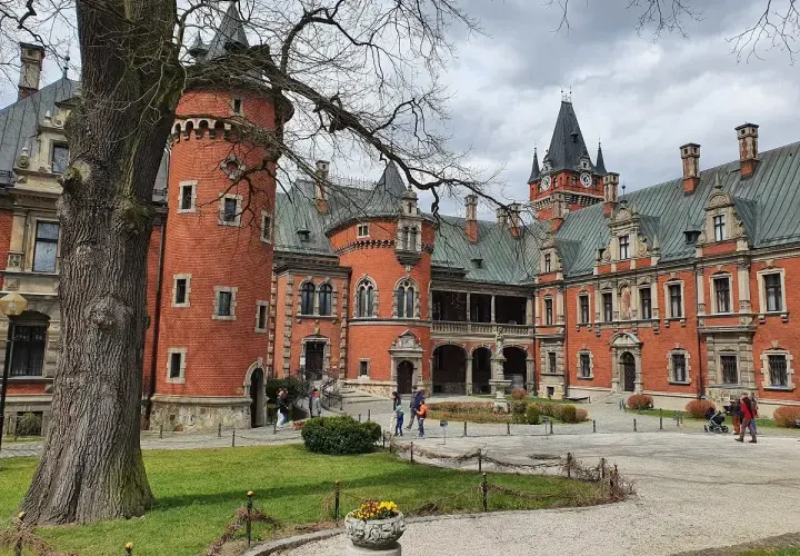 Pławniowice atrakcje: ceglany zamek z wieżą i zegarem, dziedziniec z drzewem i ludźmi spacerującymi po okolicy.