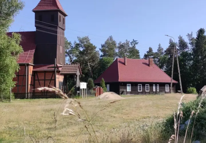 Drewniana wieża kościelna i dom z czerwonym dachem w Starowej Górze, otoczone polami i drzewami.