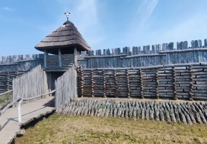 Drewniany gród obronny z wieżą i strzechą, pod niebieskim niebem.