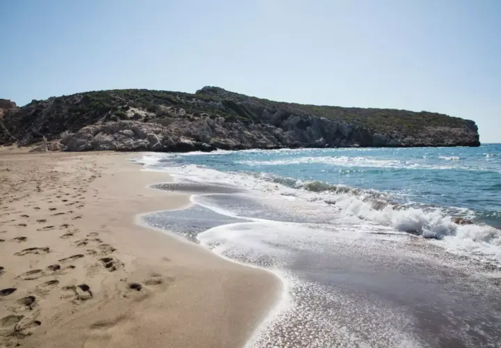 Piaszczysta plaża z falami i skalistym wzgórzem w tle.