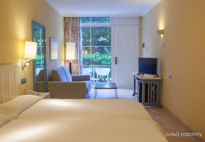 Pokój rodzinny w hotelu IFA Altamarena: łóżko, sofa, TV i wyjście na taras z widokiem na zieleń.