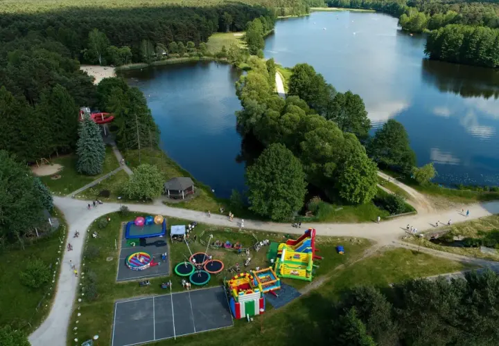 Rekreacja nad jeziorem w Bełchatowie: plac zabaw z dmuchańcami, trampolinami i zjeżdżalnią wodną, otoczony lasem i wodą.