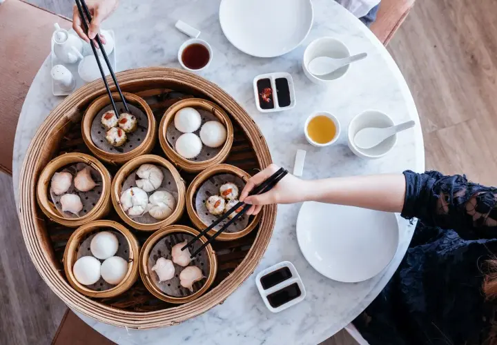 Ręce sięgają po pyszne dim sum z bambusowego parownika. Na stole stoją miseczki z sosami i herbatą.