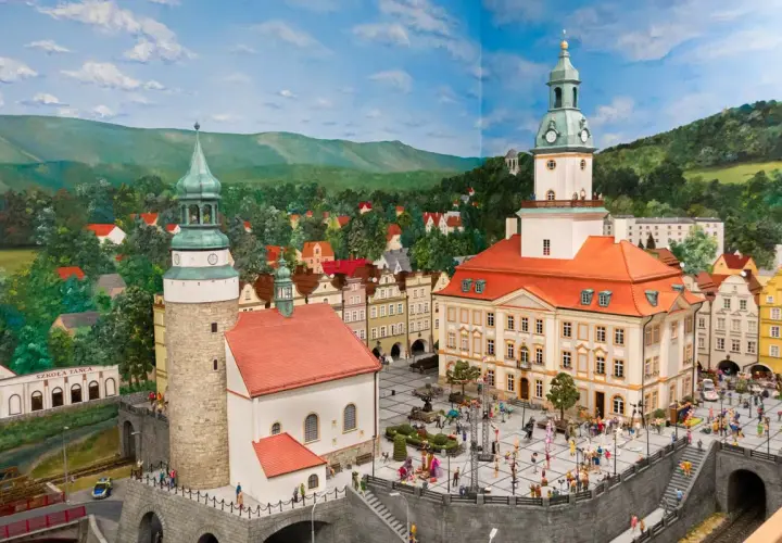 Miniaturowa panorama miasta z wieżami, ratuszem i placem pełnym postaci. W tle wzgórza i lasy, jakby z krainy jagniątków.