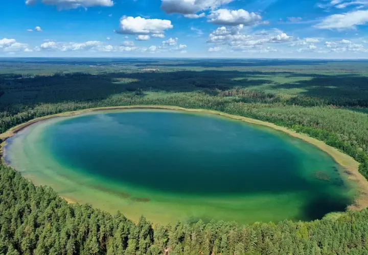 Widok z lotu ptaka na owalne, turkusowe jezioro otoczone gęstym lasem sosnowym.