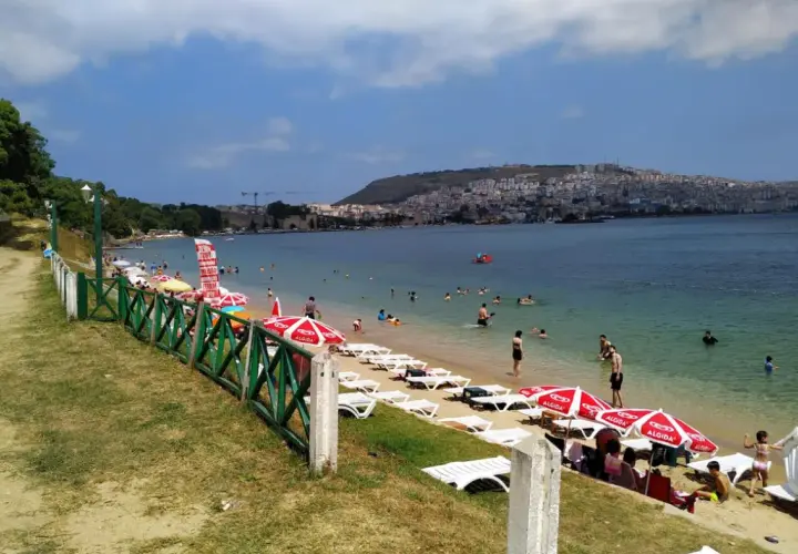 Plaża z leżakami, parasolami i ludźmi kąpiącymi się w morzu, w tle miasto na wzgórzu.