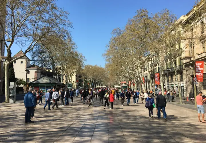Tłum ludzi spaceruje po słynnej alei **La Rambla w Barcelonie**, otoczonej drzewami i budynkami.