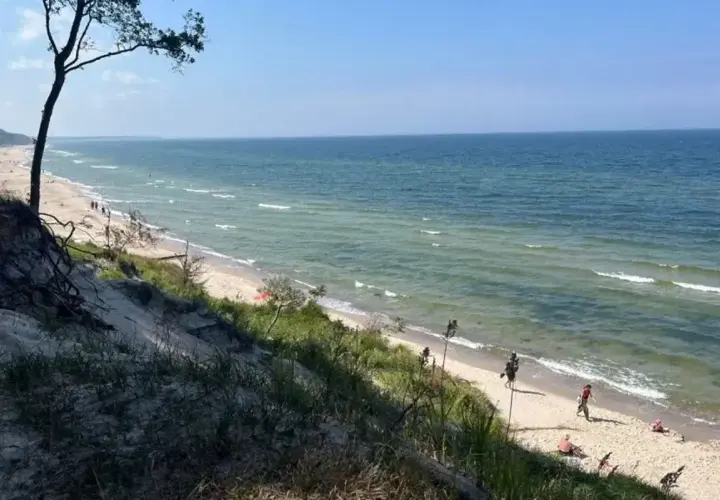 Piękna plaża z wydmami, idealna na orzechowo atrakcje. Woda w morzu jest spokojna, a na piasku widać kilka osób odpoczywających.