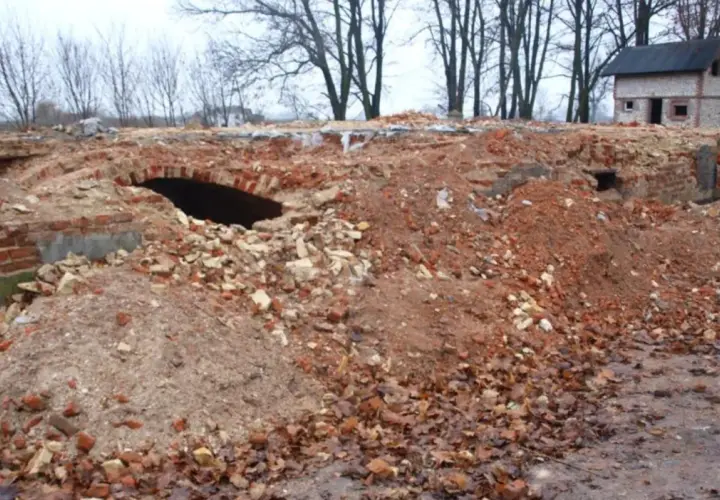 Ruiny ceglane z łukiem i mały domek w tle, otoczone drzewami.