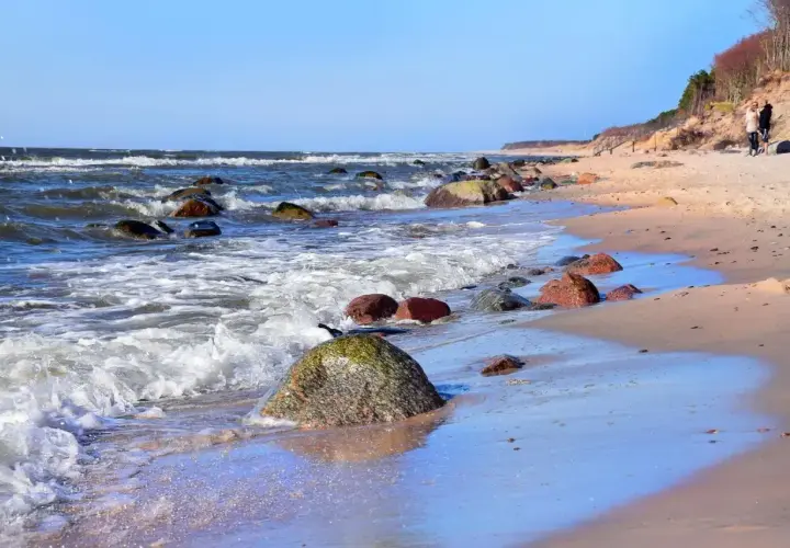 Kamienista plaża w Dębinie, z falami rozbijającymi się o brzeg i dwiema osobami spacerującymi po piasku. Dębina atrakcje przyrodnicze zachwycają.