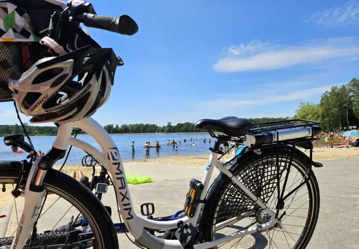 Rower z kaskiem czeka na plaży nad jeziorem, gdzie w upalny dzień kąpie się wiele osób. Idealne miejsce na wycieczkę rowerową i relaks w Puszczy Dulowskiej.