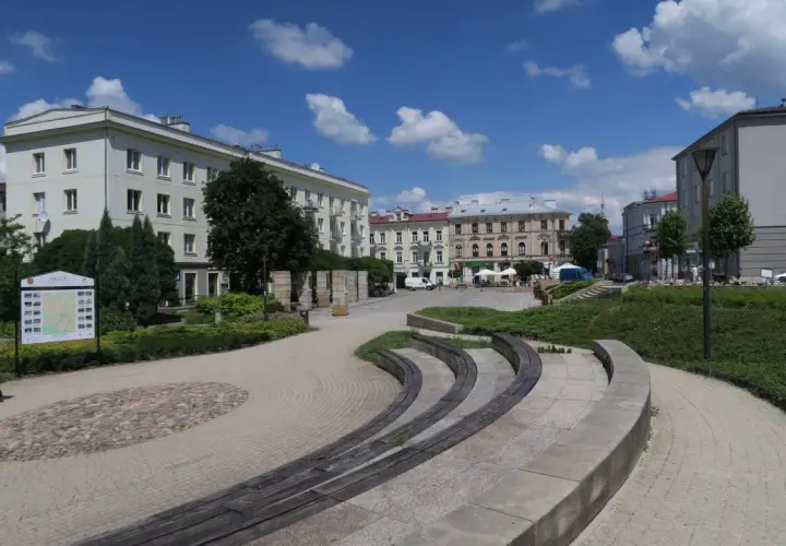 Słoneczny dzień na placu artystów w Kielcach. Widać budynki, zieleń i ławki.