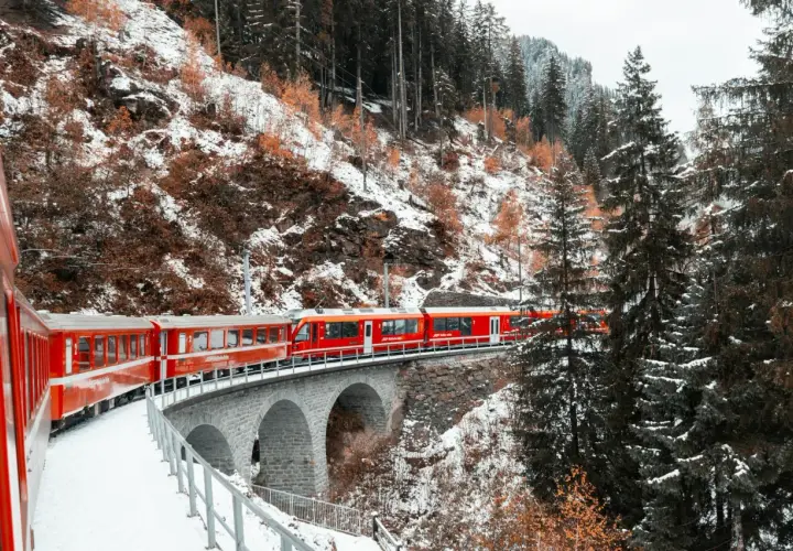Czerwony pociąg Glacier Express przejeżdża przez kamienny most w zimowym, zaśnieżonym krajobrazie górskim.