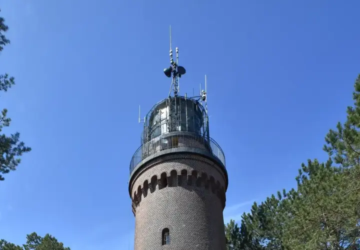 Ceglana łeba latarnia morska z metalową galeryjką i antenami na szczycie, otoczona sosnami, na tle błękitnego nieba.