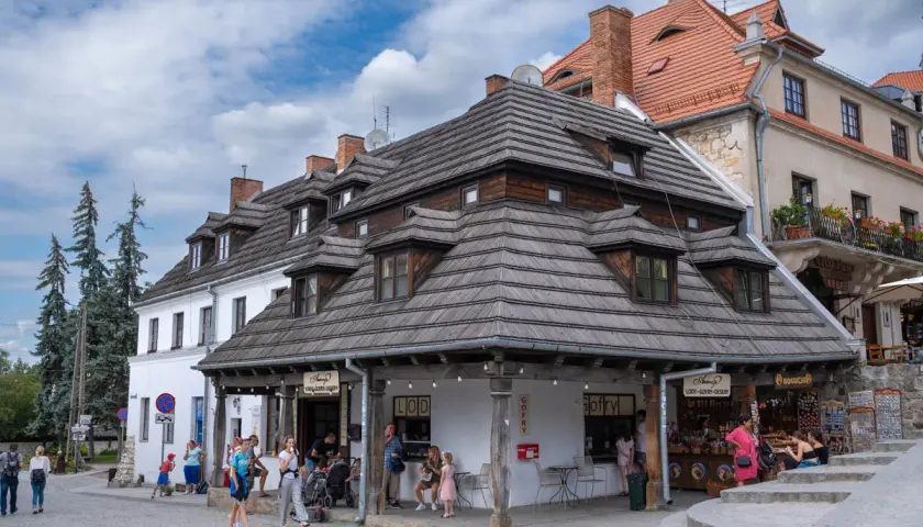 Tętniący życiem rynek w Kazimierzu Dolnym. Ludzie spacerują, kupują lody i gofry pod charakterystycznymi drewnianymi dachami.