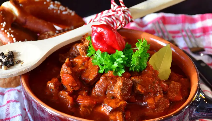 Gulasz w glinianej misie, ozdobiony papryczką i natką, z drewnianą łyżką pełną pieprzu i precelkami w tle. Idealny na chłodne wieczory, smakuje jak u babci.