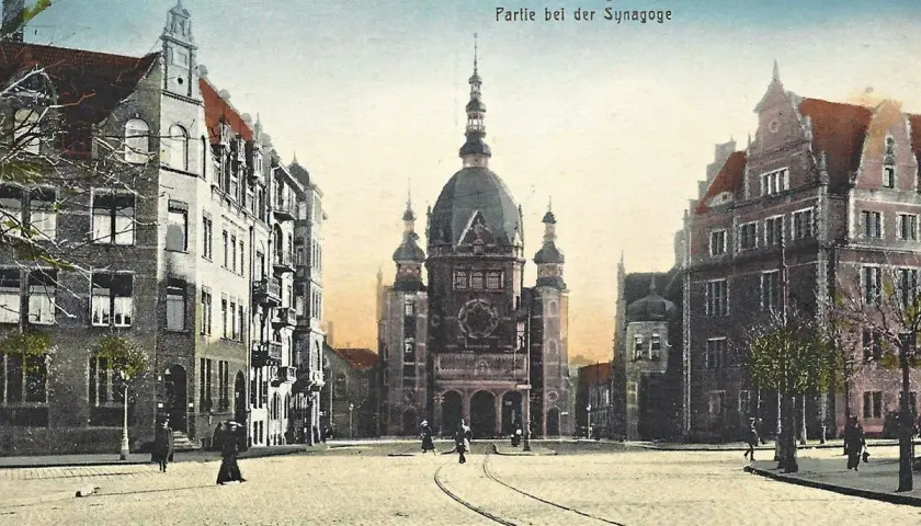 Widok na plac z imponującą synagogą w Gdańsku. Wokół kamienice, tory tramwajowe i kilka postaci spacerujących po bruku.
