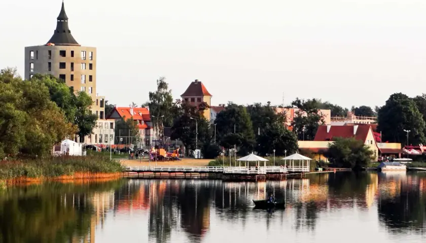 Widok na jezioro z łodzią, pomostem z altanami i budynkami w tle.