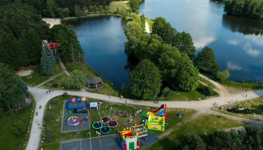 Rekreacja nad jeziorem w Bełchatowie: plac zabaw z dmuchańcami, trampolinami i zjeżdżalnią wodną, otoczony lasem i wodą.