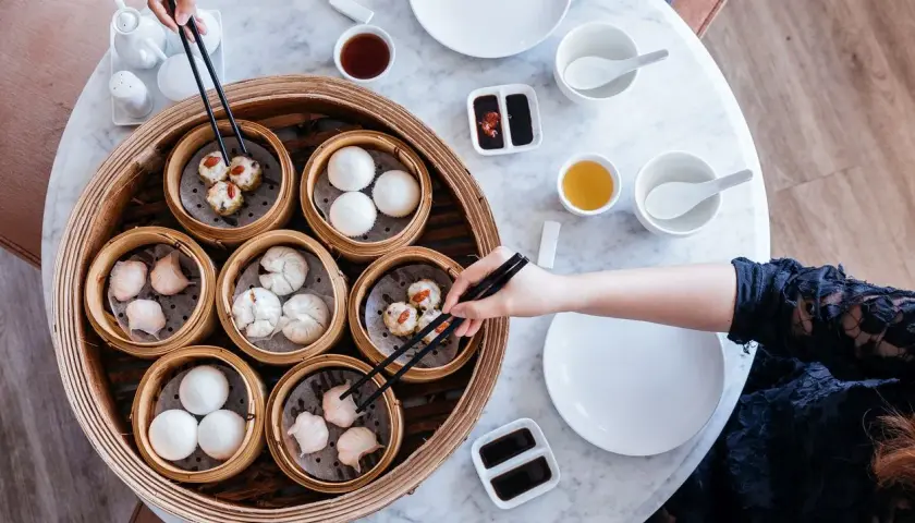 Ręce sięgają po pyszne dim sum z bambusowego parownika. Na stole stoją miseczki z sosami i herbatą.