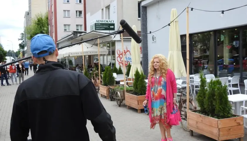 Kobieta w kolorowej sukience i różowym kardiganie czeka na ekipę filmową. W tle widać bistro i ludzi, którzy idą do stołu marsz.