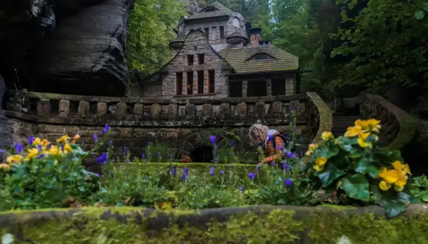 Kamienny domek w lesie, z kwitnącymi kwiatami i osobą w tle.