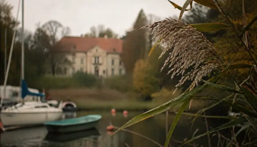 Jesienny widok na jezioro w Sztynorcie. W oddali pałac, na wodzie łodzie i żaglówka. Sztynort atrakcje dla miłośników przyrody i historii.