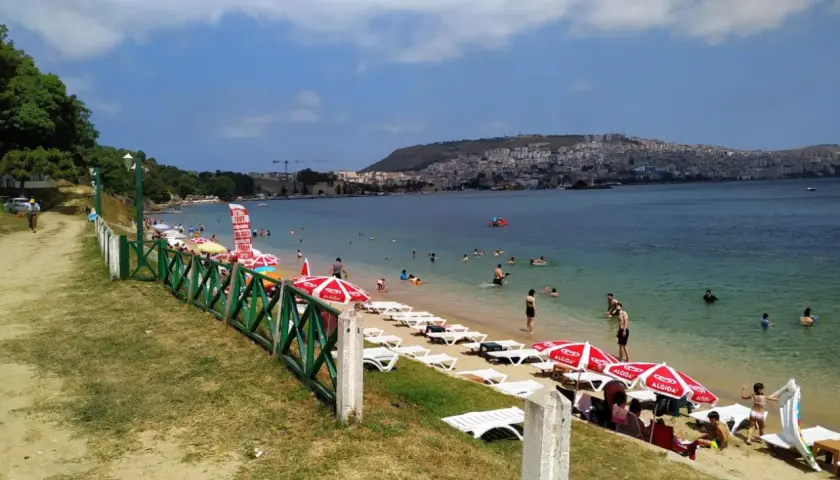 Plaża z leżakami, parasolami i ludźmi kąpiącymi się w morzu, w tle miasto na wzgórzu.