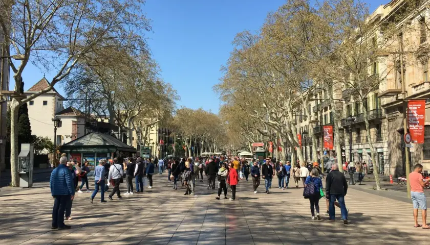 Tłum ludzi spaceruje po słynnej alei **La Rambla w Barcelonie**, otoczonej drzewami i budynkami.
