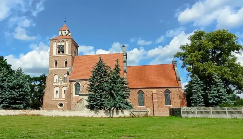 Nieszawa atrakcje: ceglany kościół z wieżą i zegarem, otoczony zielenią, na tle błękitnego nieba z chmurami.